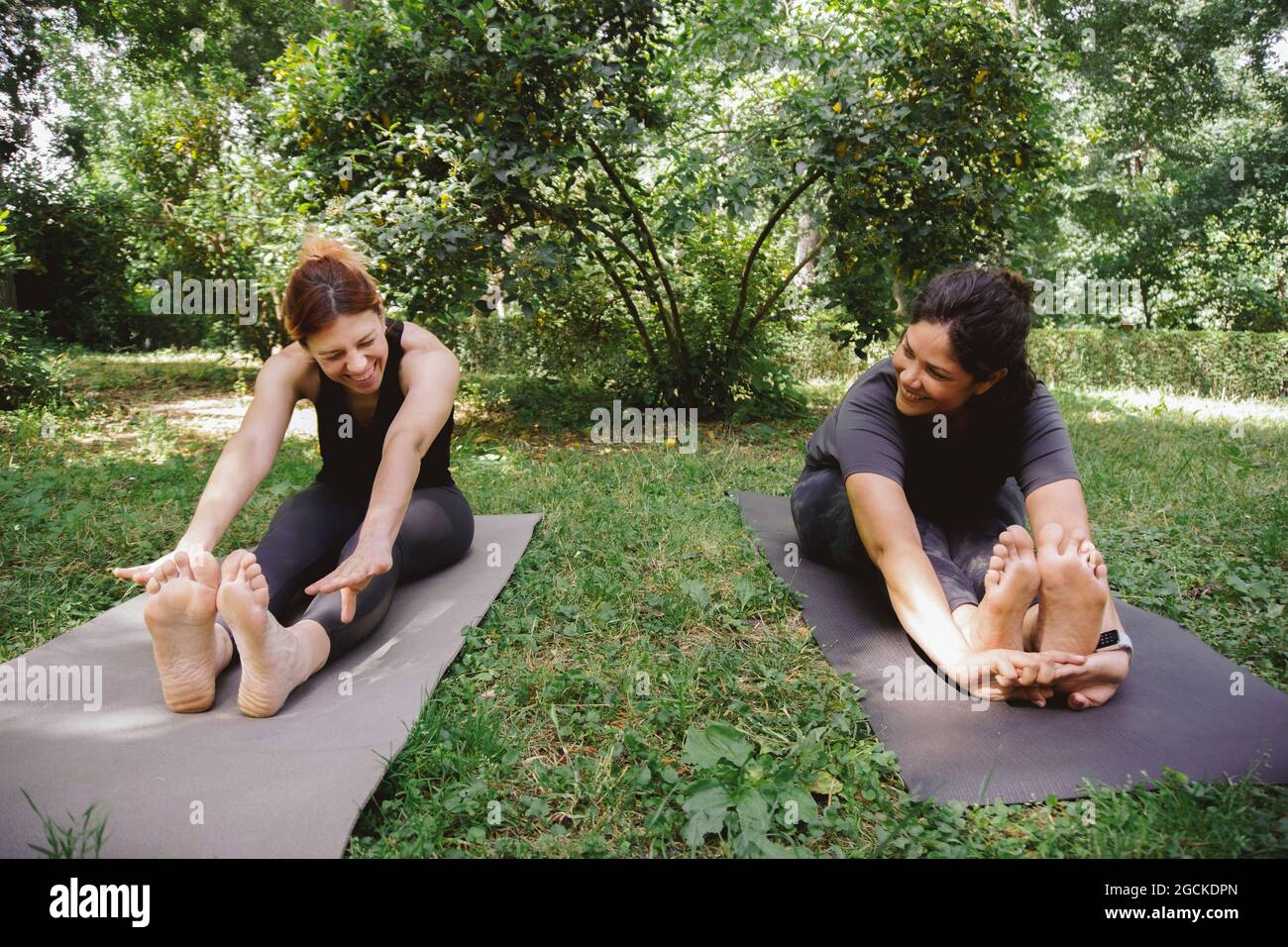 Full body of positive young Woman in activewear sitting on mat on lawn smiling while trying to perform Seated Forward Bend I asana during yoga session Stock Photo