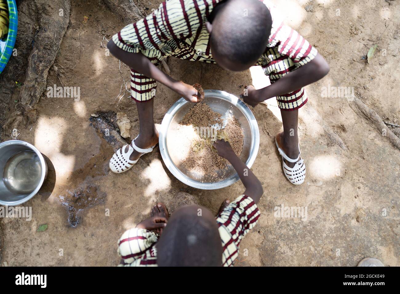 In this image two small black African brothers in identical clothes are sharing a meat-free plate of rice, sitting on the ground, with their right han Stock Photo