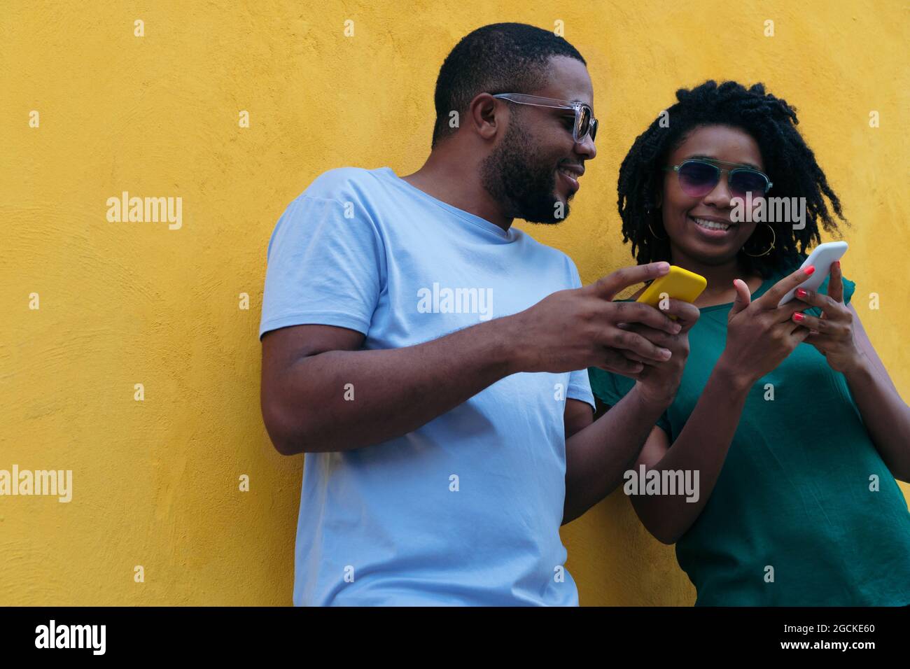 Black couple using a mobile phone leaning against a yellow wall Stock Photo