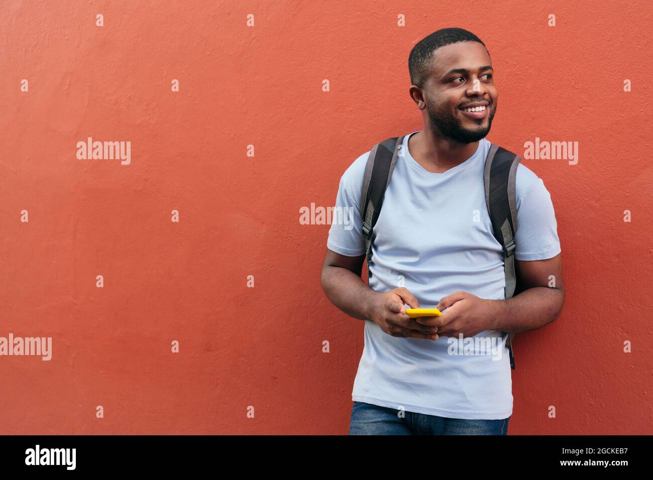 African man with backpack and mobile phone smiling while leaning on wall Stock Photo