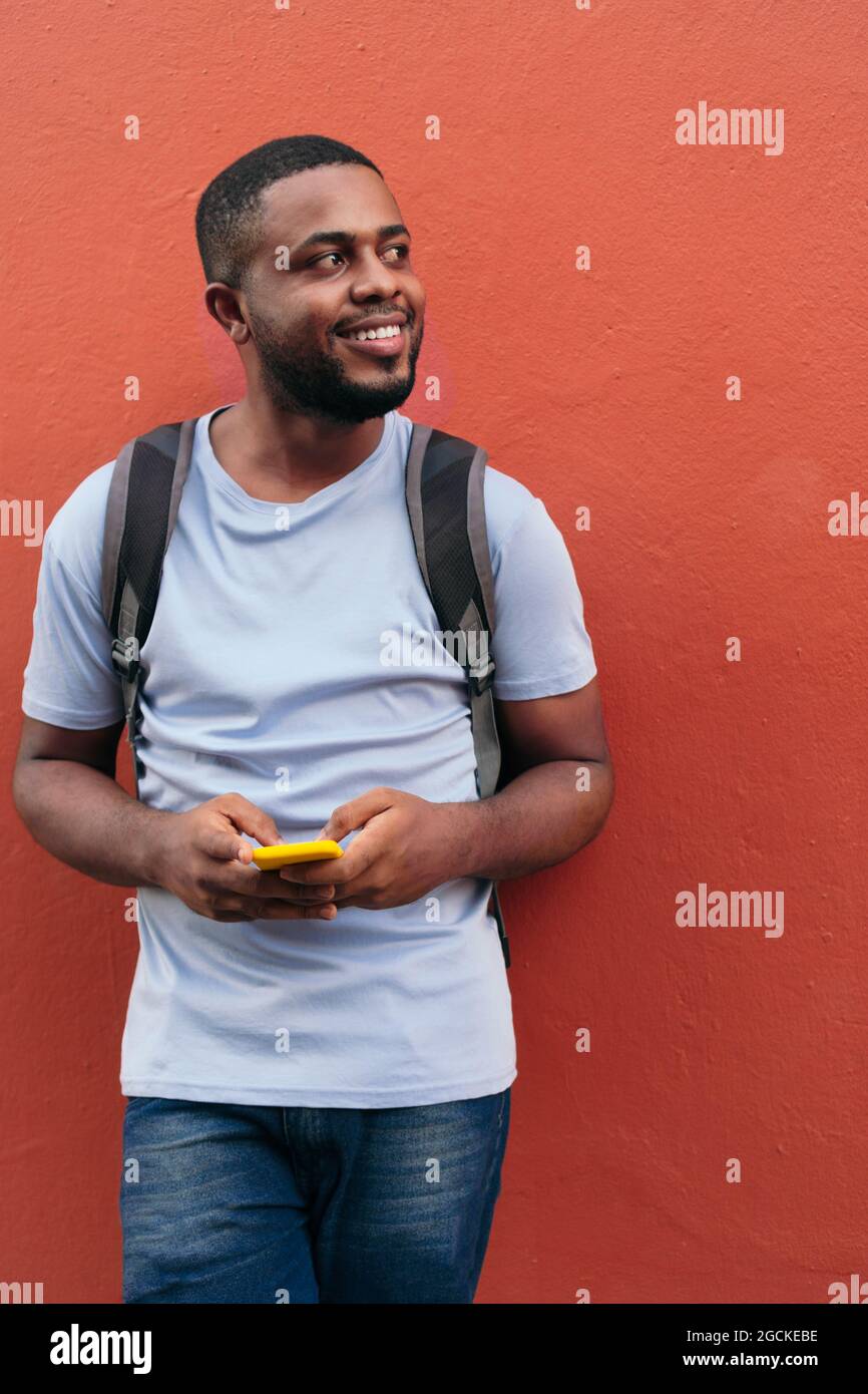 Black man with backpack and mobile phone smiling while leaning on wall Stock Photo