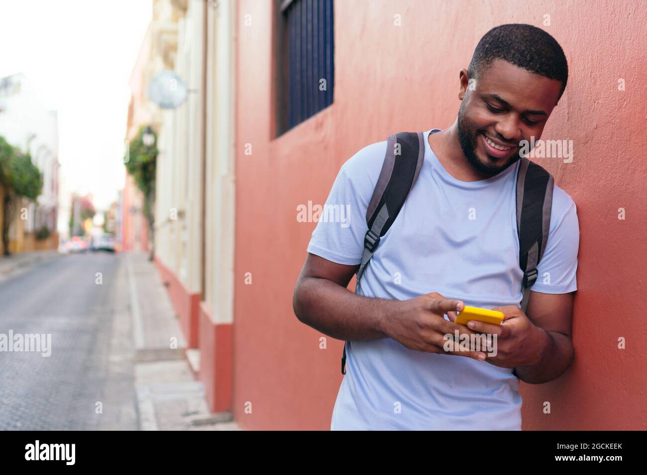 man with backpack checking cell phone while leaning against wall Stock Photo
