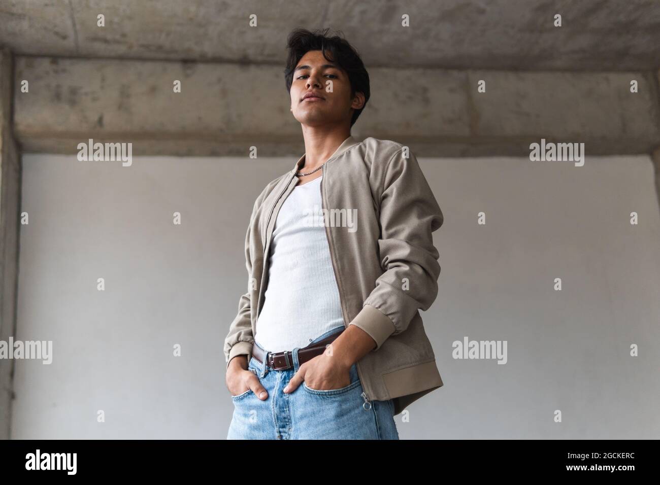From below portrait of young latin man looking confidently at camera with hands on pockets against neutral background Stock Photo