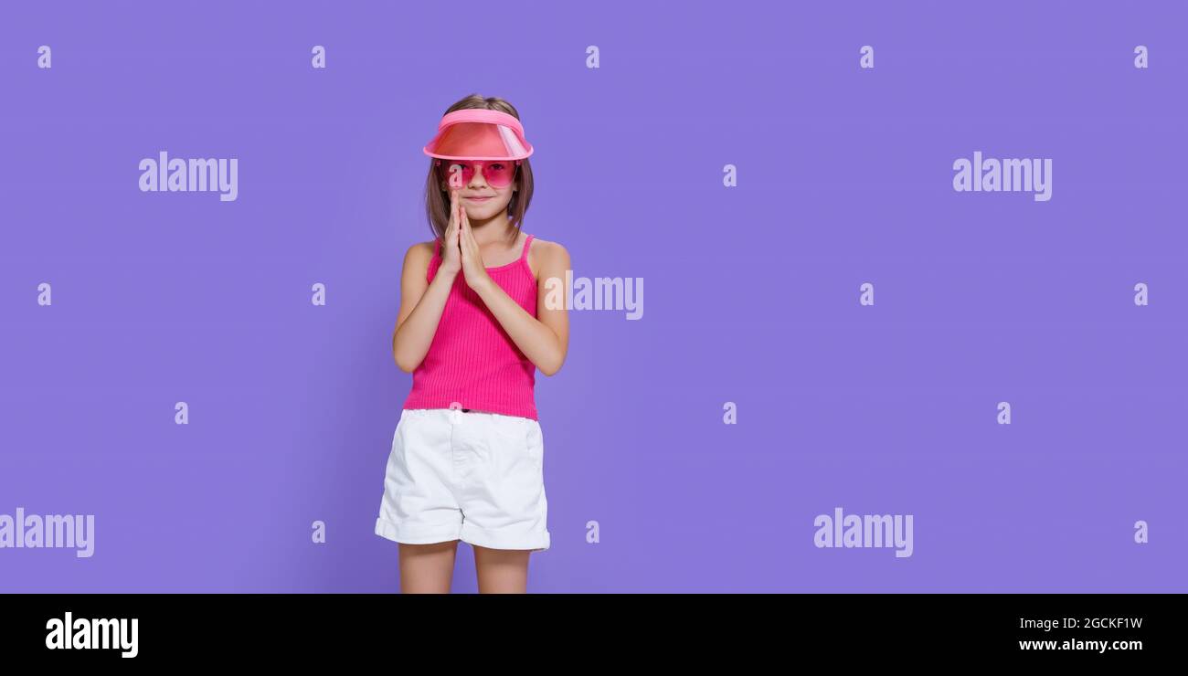 a little girl in white shorts, a pink T-shirt, pink trendy glasses and a summer visor on a purple background in the studio Stock Photo