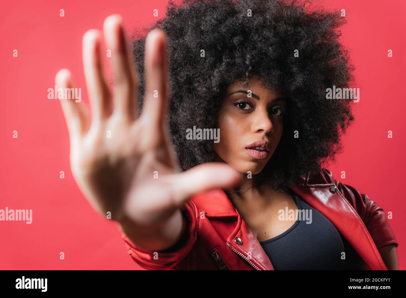 Frightened African American female with curly hair showing stop gesture on red background in studio and looking at camera Stock Photo