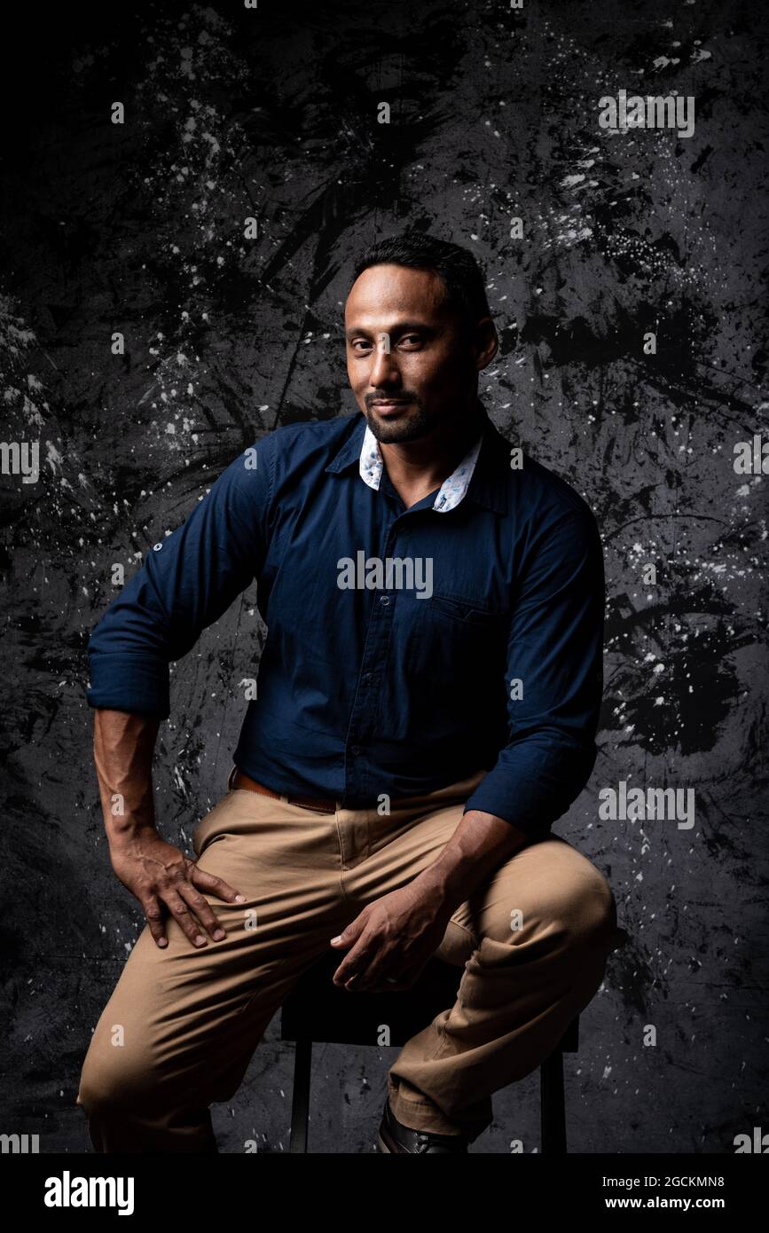 Serious stylish ethnic male sitting on wooden stool in studio on dark background and looking at camera Stock Photo