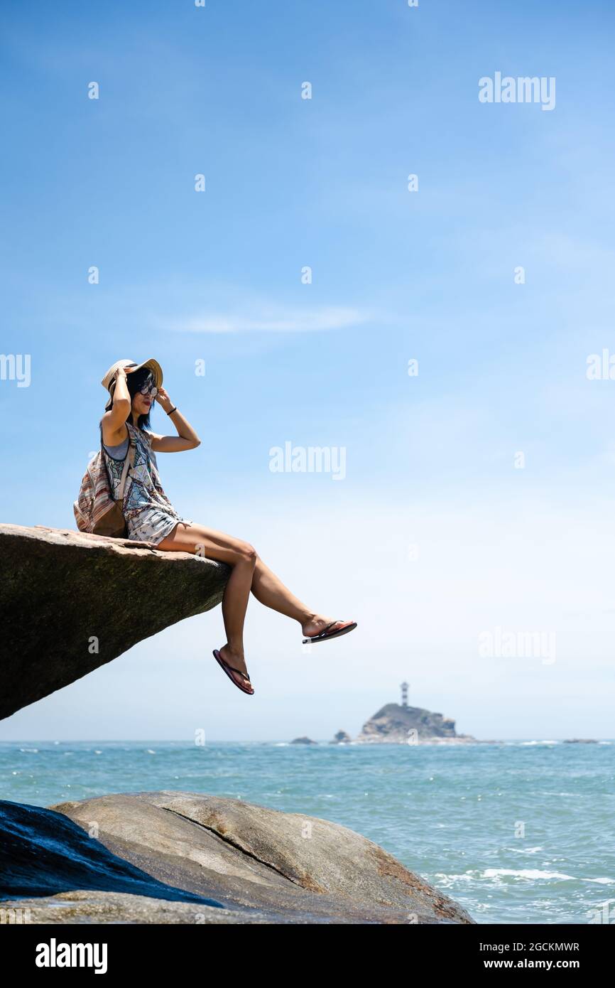 Beautiful Asian girl sitting on a rock, enjoying the seaside in sunny summer's day Stock Photo