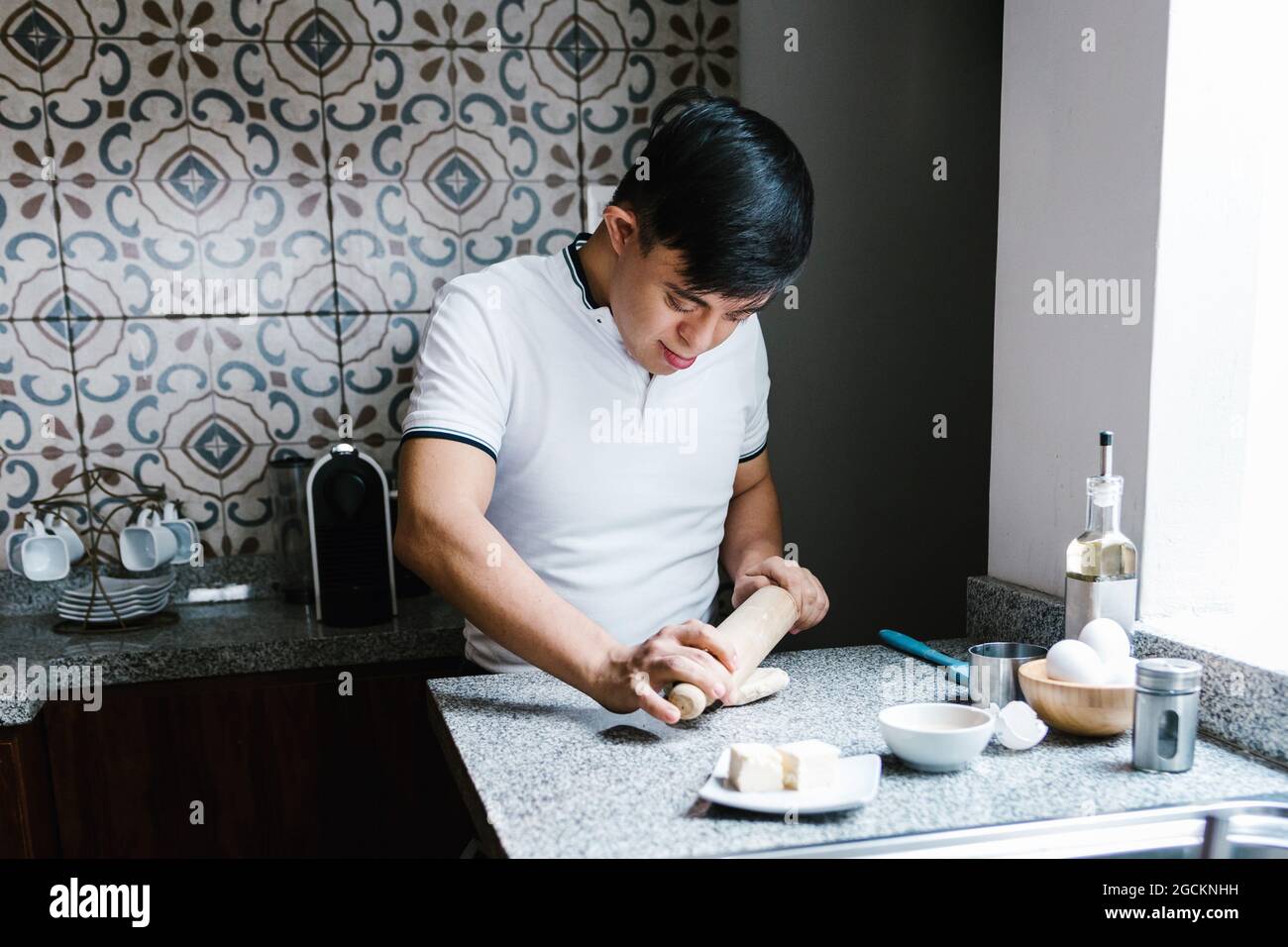 Focus ethnic teenage boy with Down syndrome rolling dough with rolling pin while cooking in kitchen Stock Photo