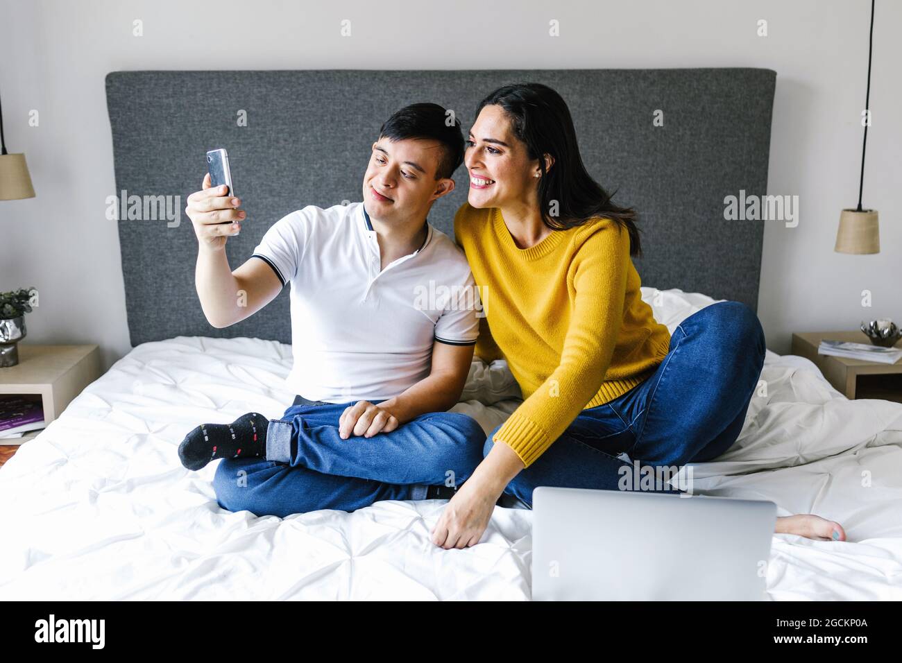 Delighted ethnic mother and teenage boy with Down syndrome sitting on bed and taking self portrait on smartphone Stock Photo
