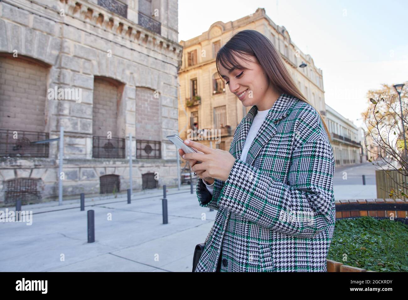 Cheerful young female in stylish houndstooth coat using mobile phone while standing on urban square Stock Photo