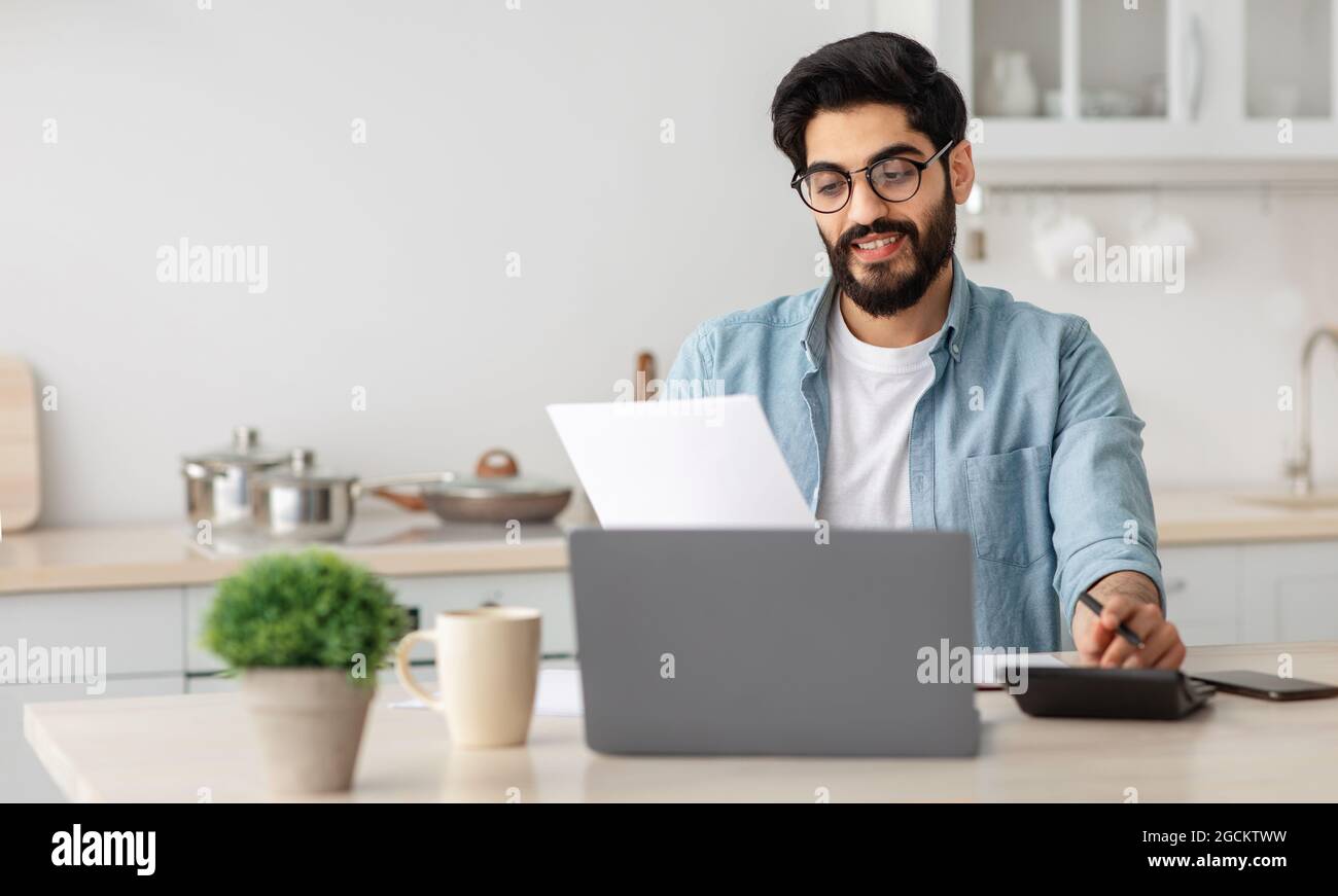 Portrait of happy arab guy reading insurance documents at home, sitting at table in kitchen and counting home budget Stock Photo