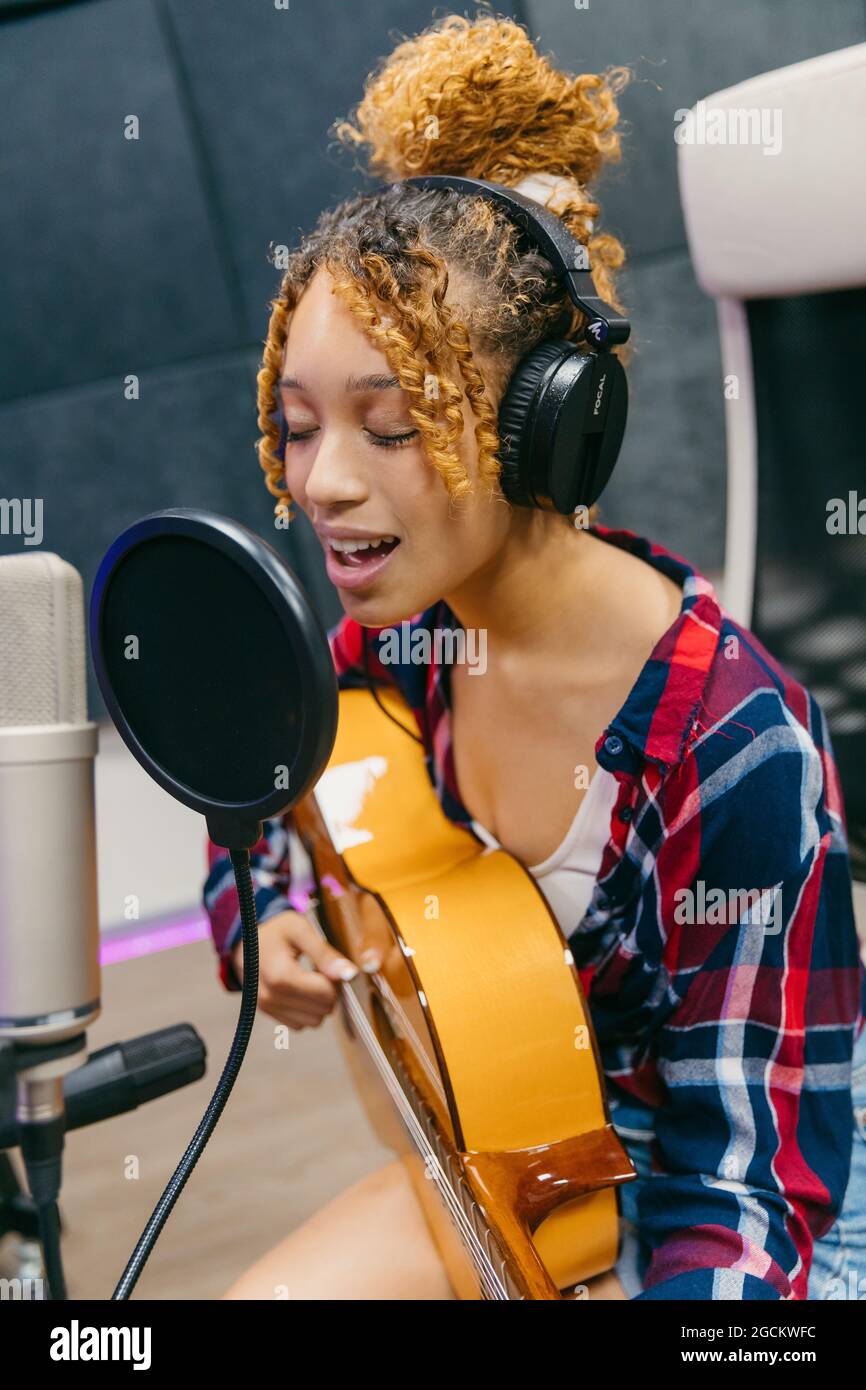 Young ethnic female vocalist in headphones playing acoustic guitar