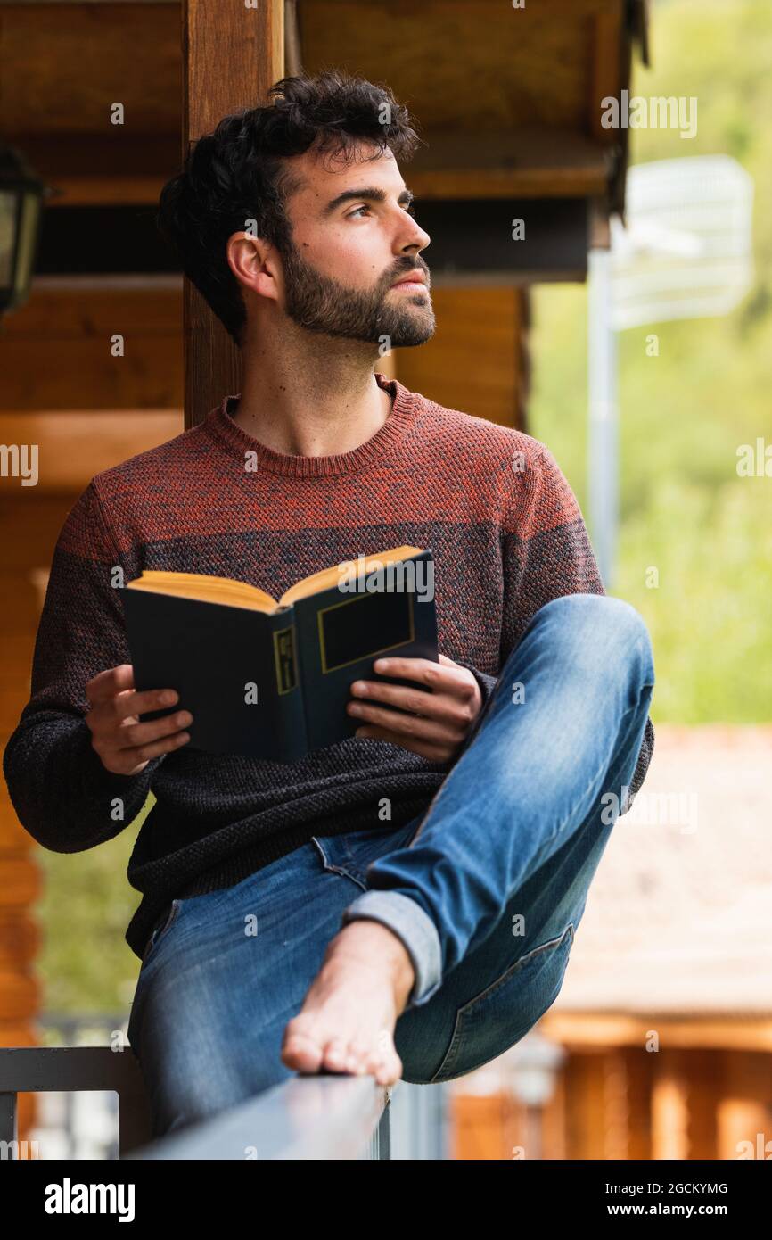 Contemplating male sitting on wooden terrace of house with book and looking away while enjoying vacation in Pyrenees Stock Photo
