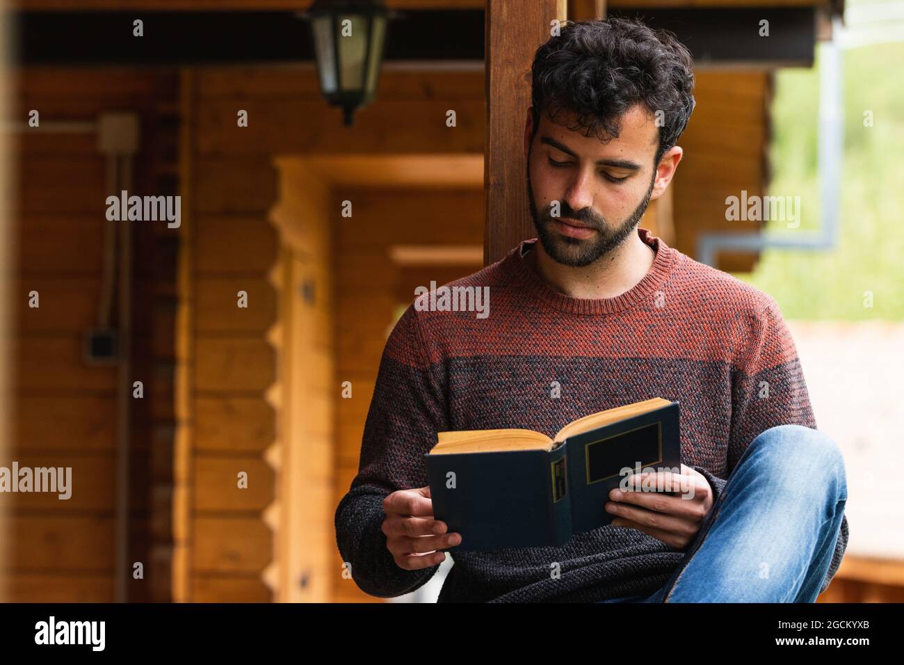Contemplating male sitting on wooden terrace of house with book and looking away while enjoying vacation in Pyrenees Stock Photo