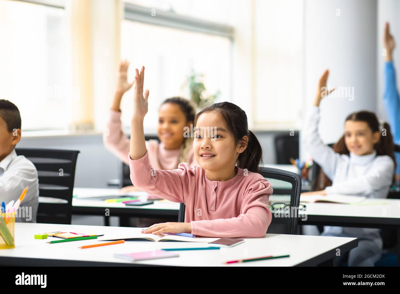Diverse group of little school children raising hands at classroom Stock Photo