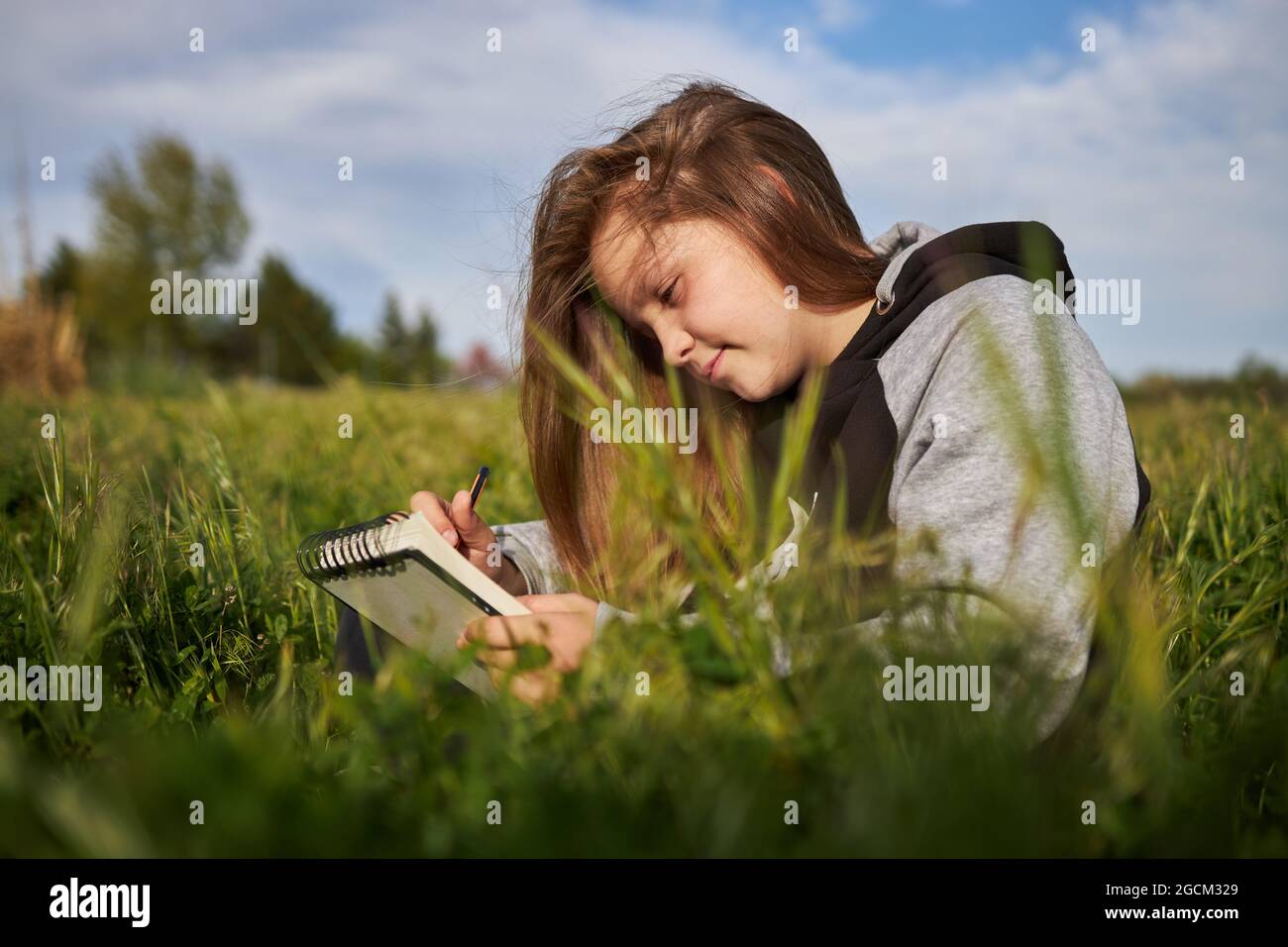 Delighted teen girl sitting in meadow and drawing in sketchbook