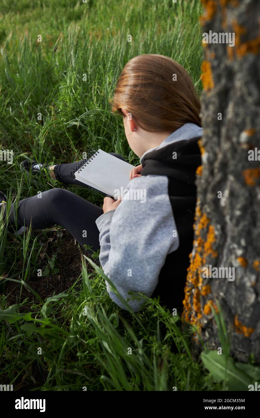 https://c8.alamy.com/comp/2GCM35M/back-view-of-unrecognizable-teen-girl-sitting-in-meadow-and-drawing-in-sketchbook-while-enjoying-sunny-day-in-countryside-leaning-on-tree-trunk-2GCM35M.jpg