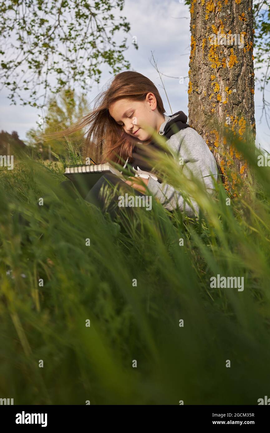 https://c8.alamy.com/comp/2GCM35R/delighted-teen-girl-sitting-in-meadow-and-drawing-in-sketchbook-while-enjoying-sunny-day-in-countryside-leaning-on-tree-trunk-2GCM35R.jpg