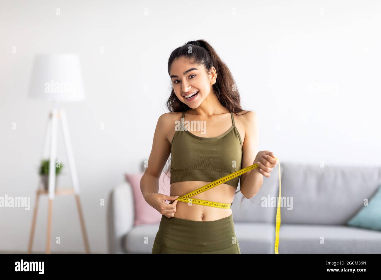 Successful weight loss. Slim Indian woman measuring waist with tape, satisfied with slimming program at home Stock Photo