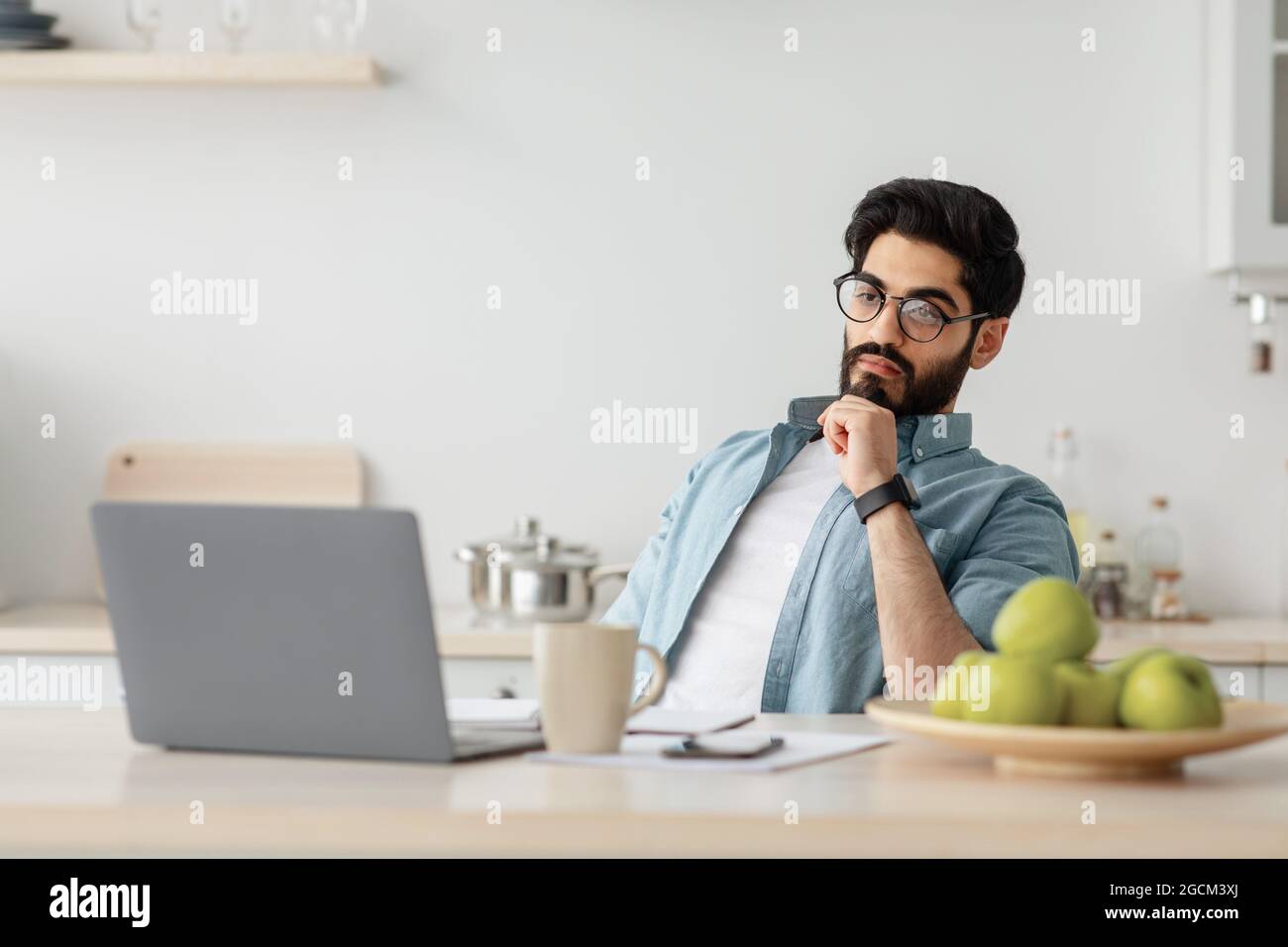 Thoughtful arab freelancer man looking at laptop screen while working remotely at home, sitting at kitchen with computer Stock Photo