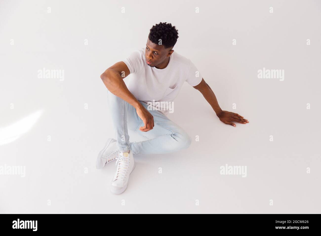 From above full body of young African American male model wearing white t shirt with light blue jeans and sneakers sitting on floor in studio Stock Photo