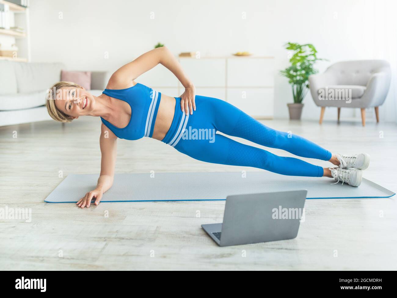Middle-aged woman doing side plank on mat with pc Stock Photo