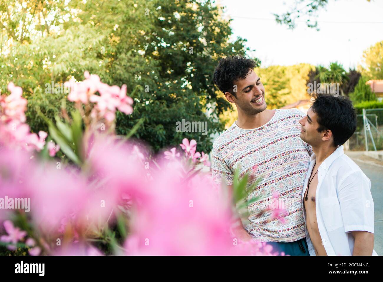 Young gay couple in love in nature with flowers in foreground. Two young gay men outdoors in summer on a street. Happy Caucasian couple in love on a d Stock Photo