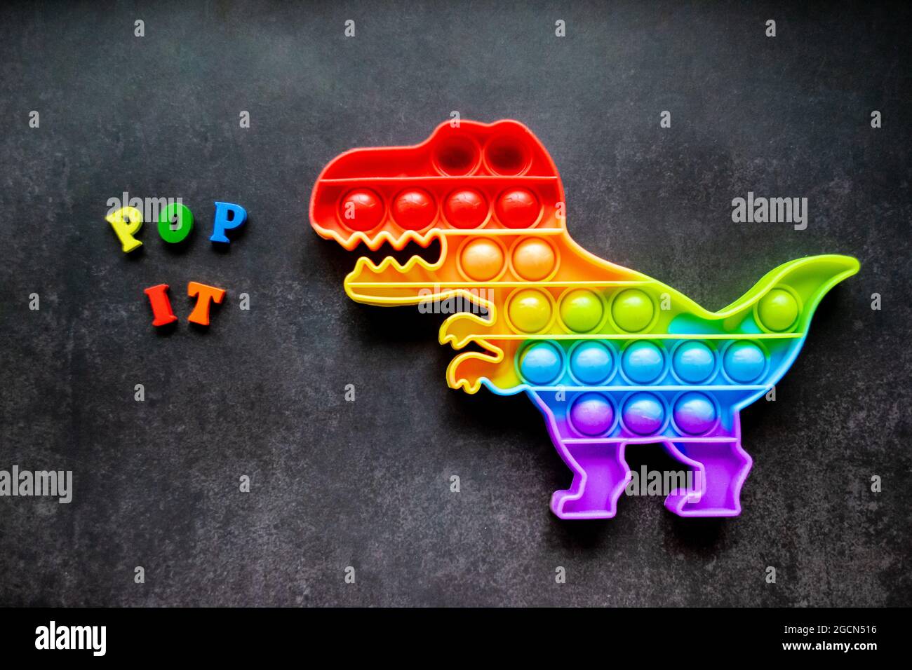 Toy pop it dinosaur rainbow colors on a black background with multicolored letters and lettering - Pop it. Stock Photo