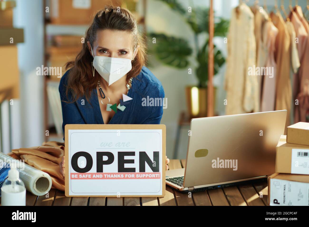trendy middle aged small business owner woman with ffp2 mask and open after covid sign in the office. Stock Photo