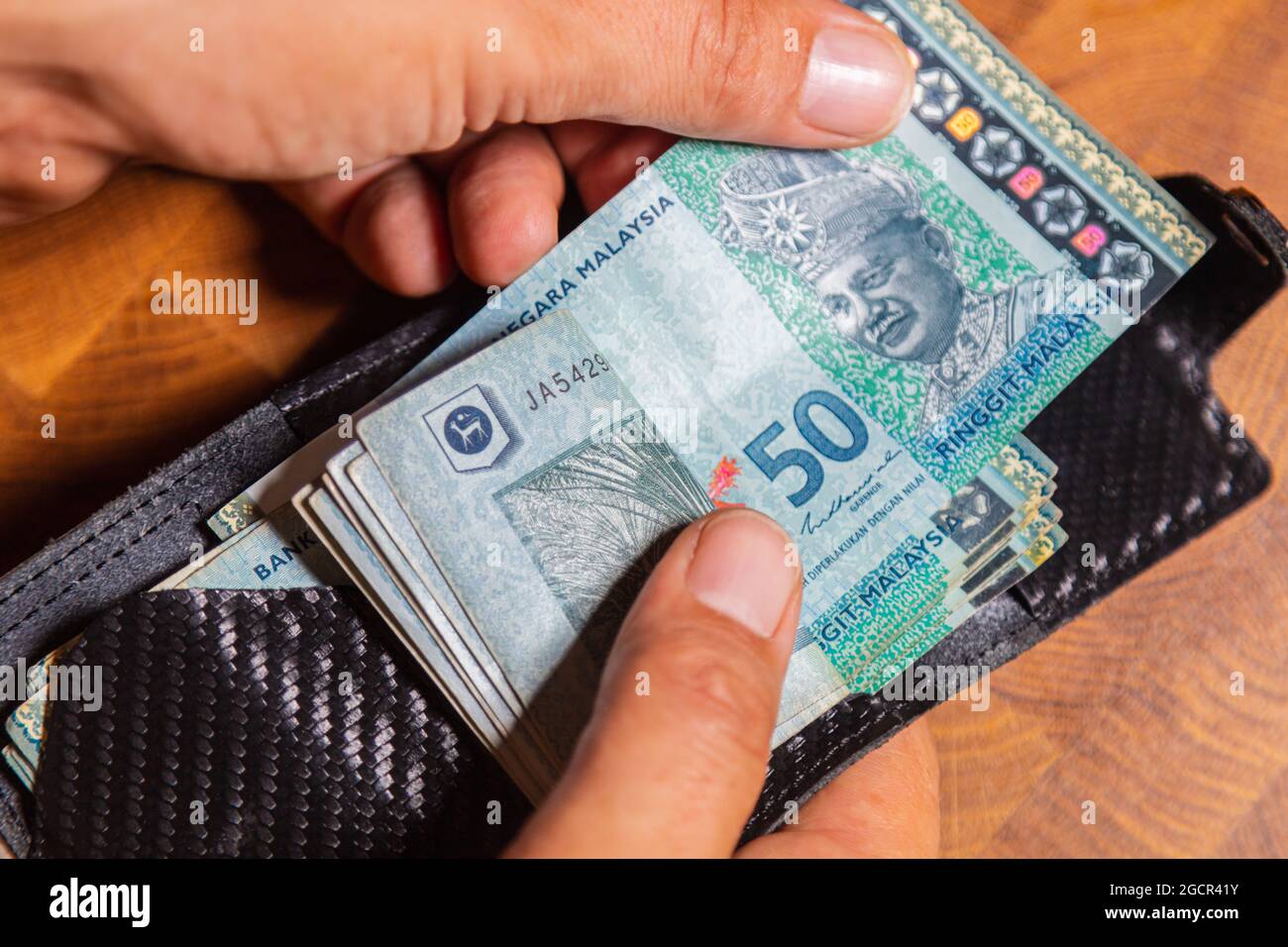 Male hands counting 50 Ringgit Malaysia in a leather wallet on a wooden table. RM50 in mans hand. Malaysian Ringgit shown by young man in a wallet. Cl Stock Photo