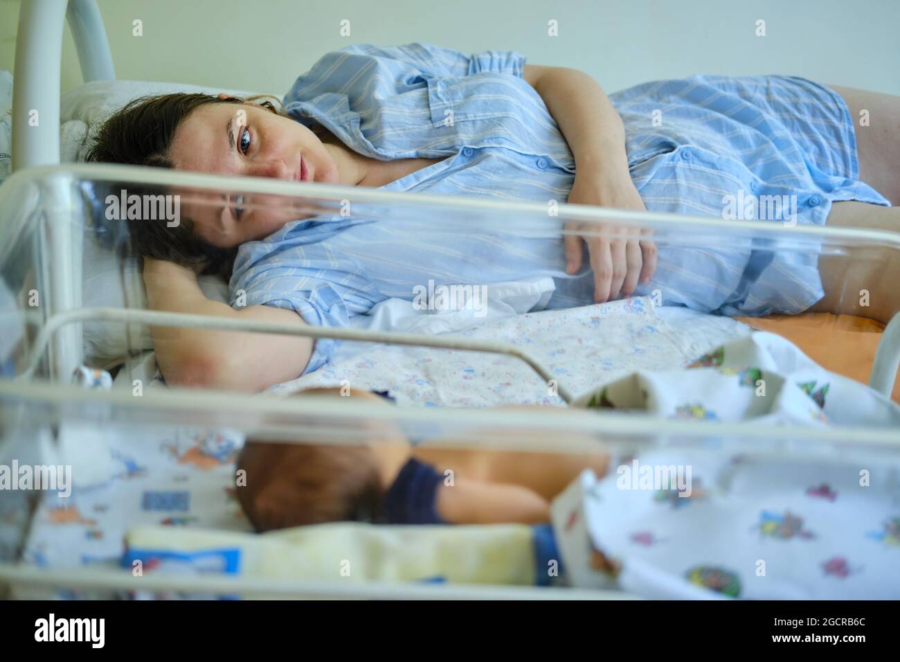 Mom is sleeping in the ward with a newborn baby, a maternity hospital bracelet on her arm. A newly born child in a clinic crib behind a transparent gl Stock Photo