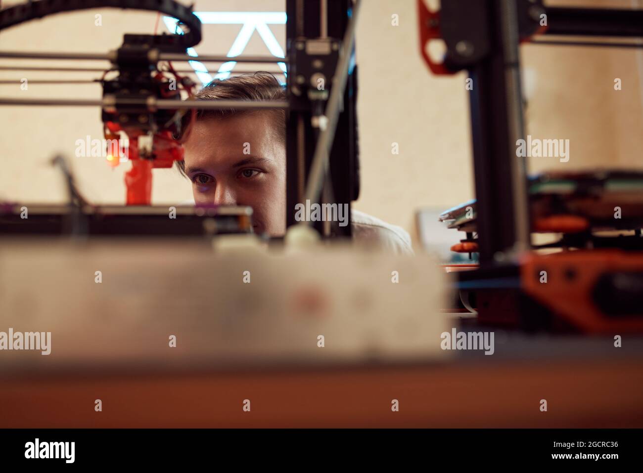 Focused young man watching modern 3D printer creating red plastic model in studio Stock Photo