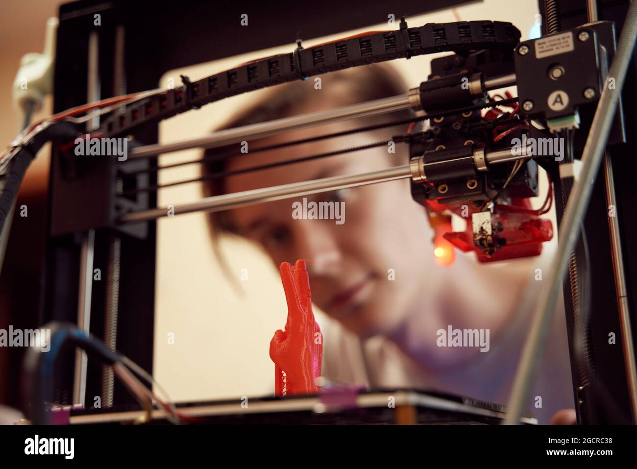 Focused young man watching modern 3D printer creating red plastic model in studio Stock Photo