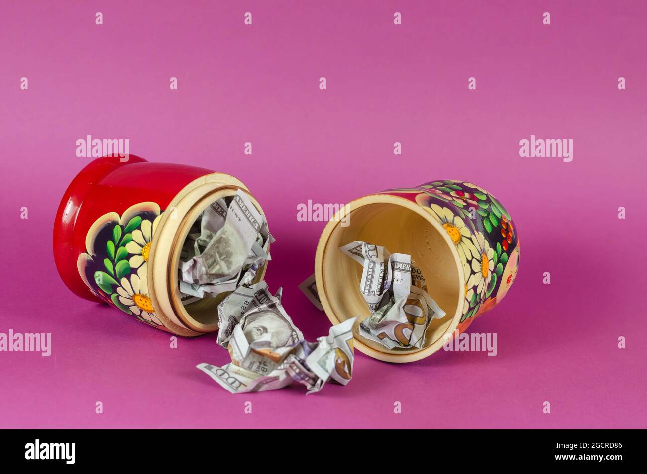 xRussian matryoshka full of money. Crumpled hundred-dollar bills fall out of open wooden toy. Symbol of Russia and American money on  pink background Stock Photo