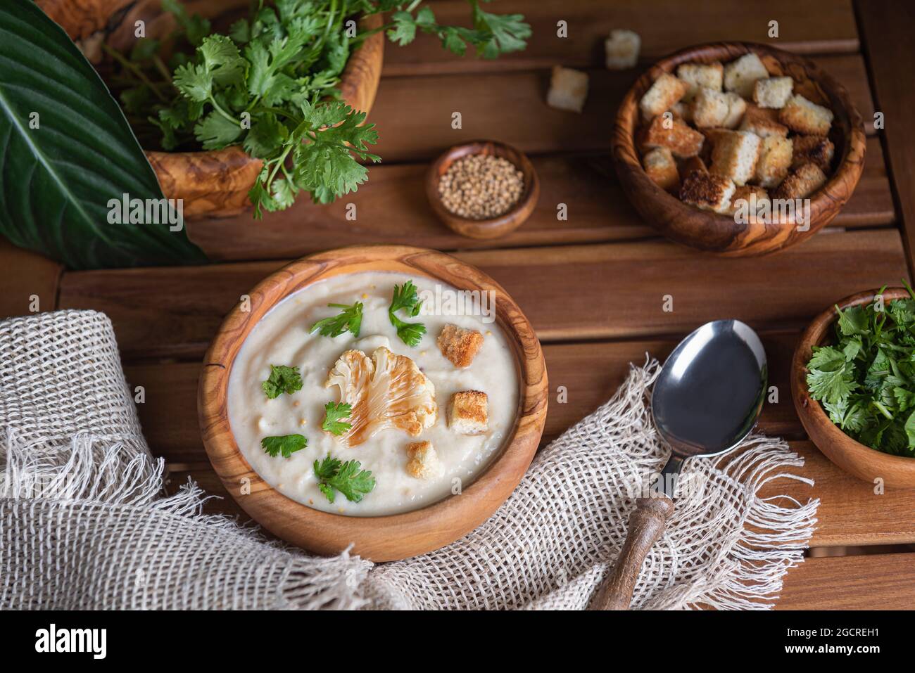 Homemade cauliflower cream soup with freshly chopped herbs and croutons in wooden bowls on brown wooden table. Linen napkin, spoon, mustard seeds and Stock Photo