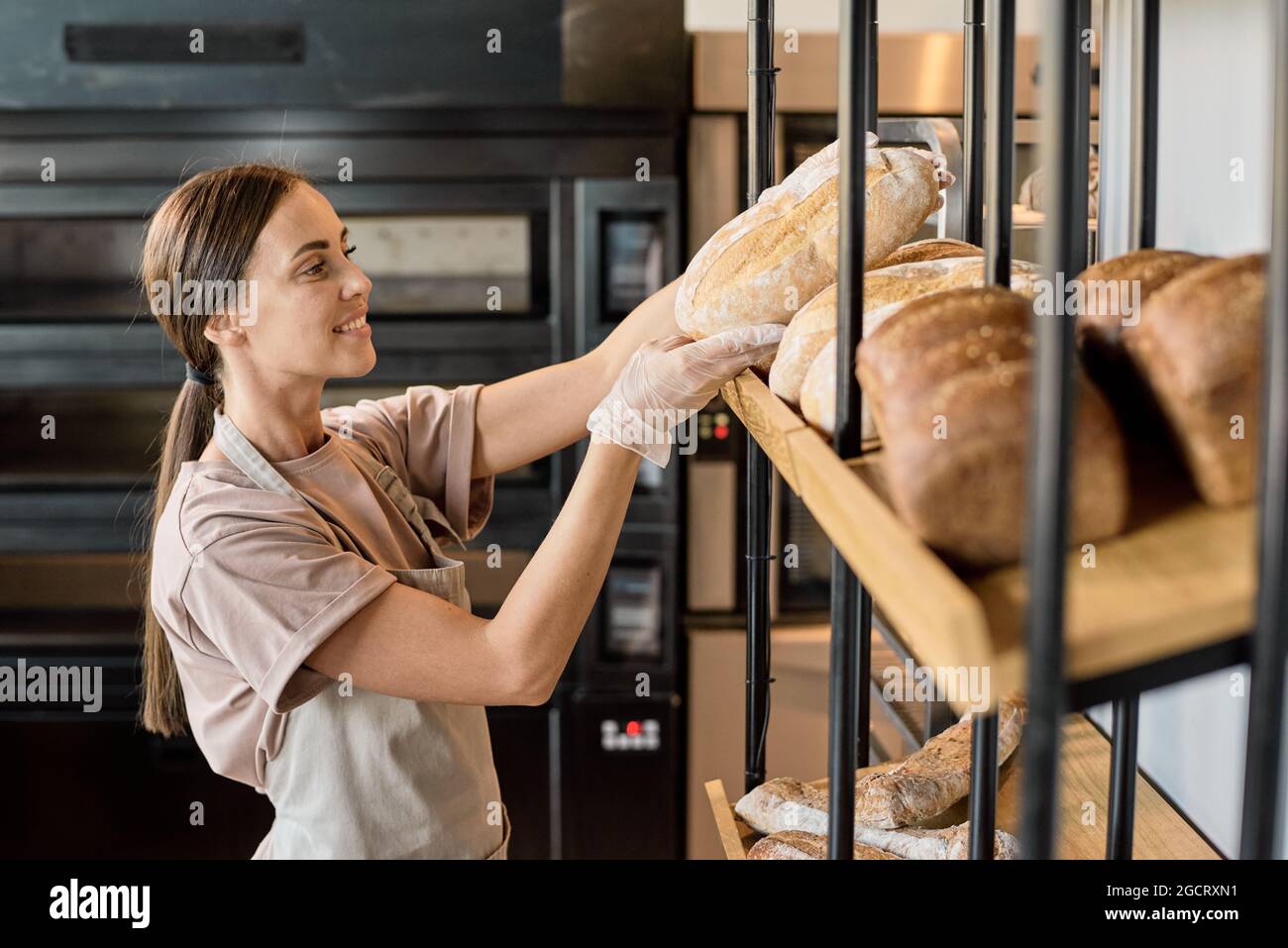 Young baker in apron putting fresh bread on display while working in