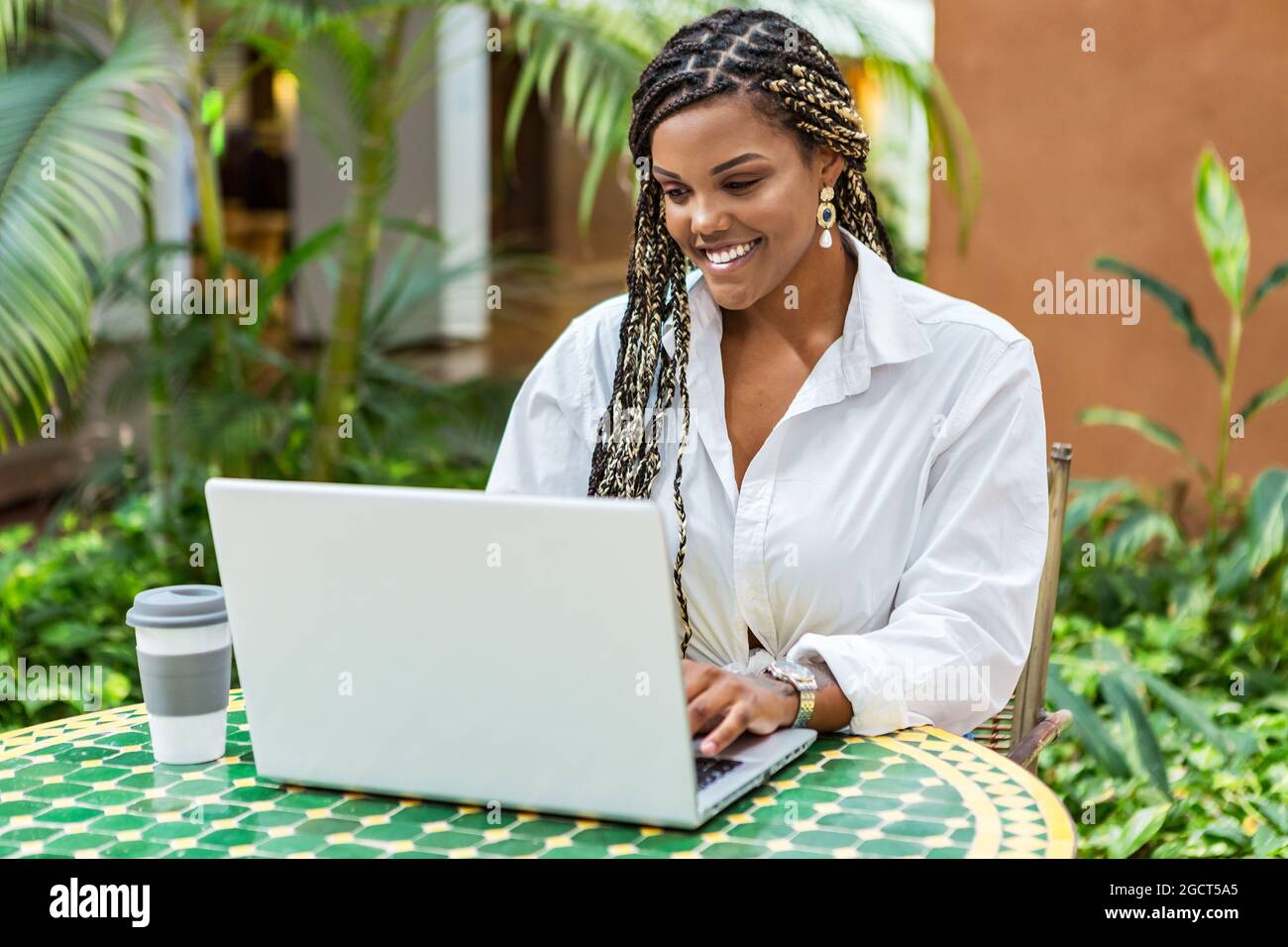 African American woman using laptop in a cafe store outside. Woman with braids sitting using laptop and drinking coffee. Stock Photo