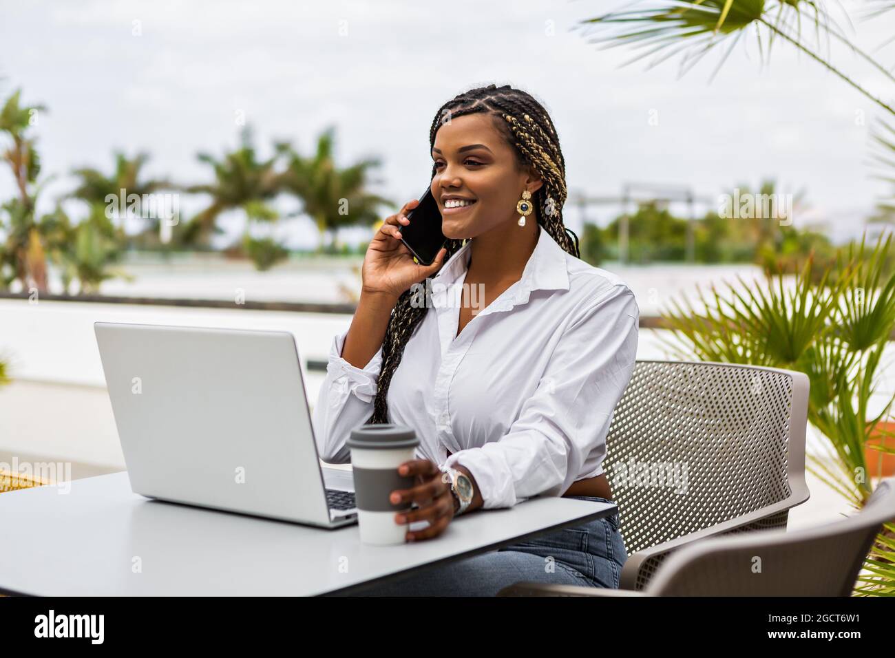 Cheerful African American woman with her laptop talking with cellphone while holding cup of coffee in a cafe outside. Woman with braids sitting using Stock Photo