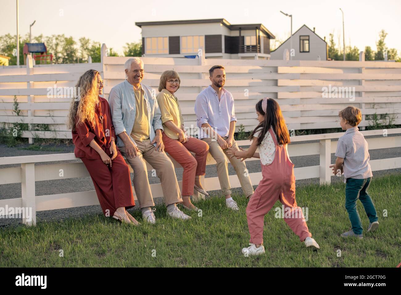 Happy young and senior couples sitting on white fence in front of two kids playing on green lawn Stock Photo