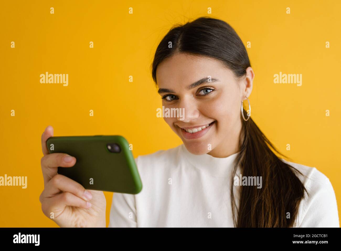 Young brunette woman smiling and using mobile phone isolated over yellow background Stock Photo
