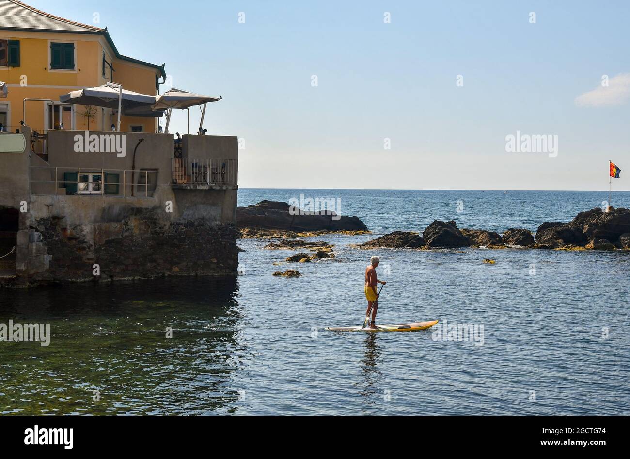 Senior man stand up paddle boarding (SUP) on the shore of the fishing village with a terrace restaurant in the background, Boccadasse, Genoa, Liguria Stock Photo