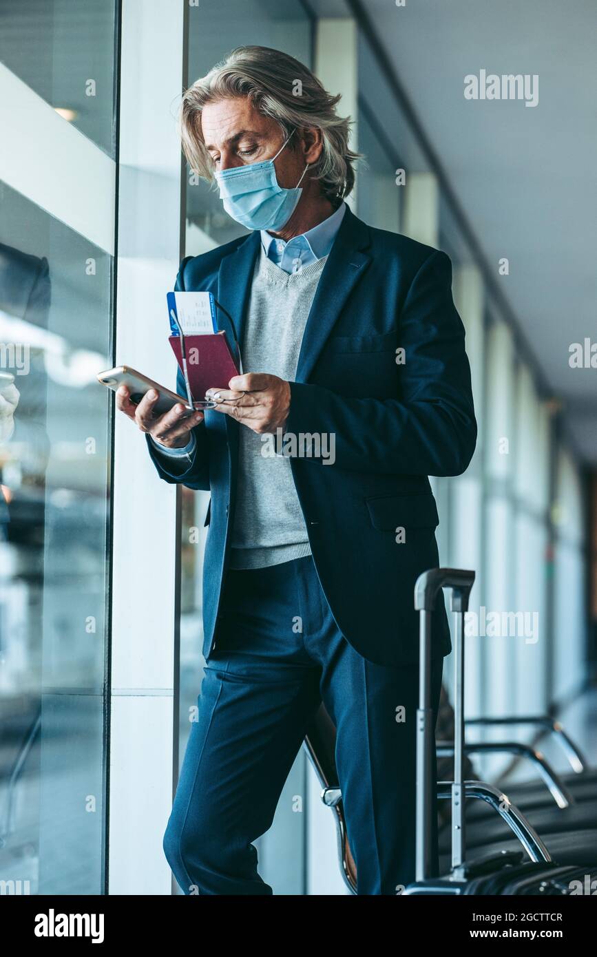 Man with face mask standing at airport waiting area using his mobile phone. traveler waiting for his flight at airport departure area. Stock Photo