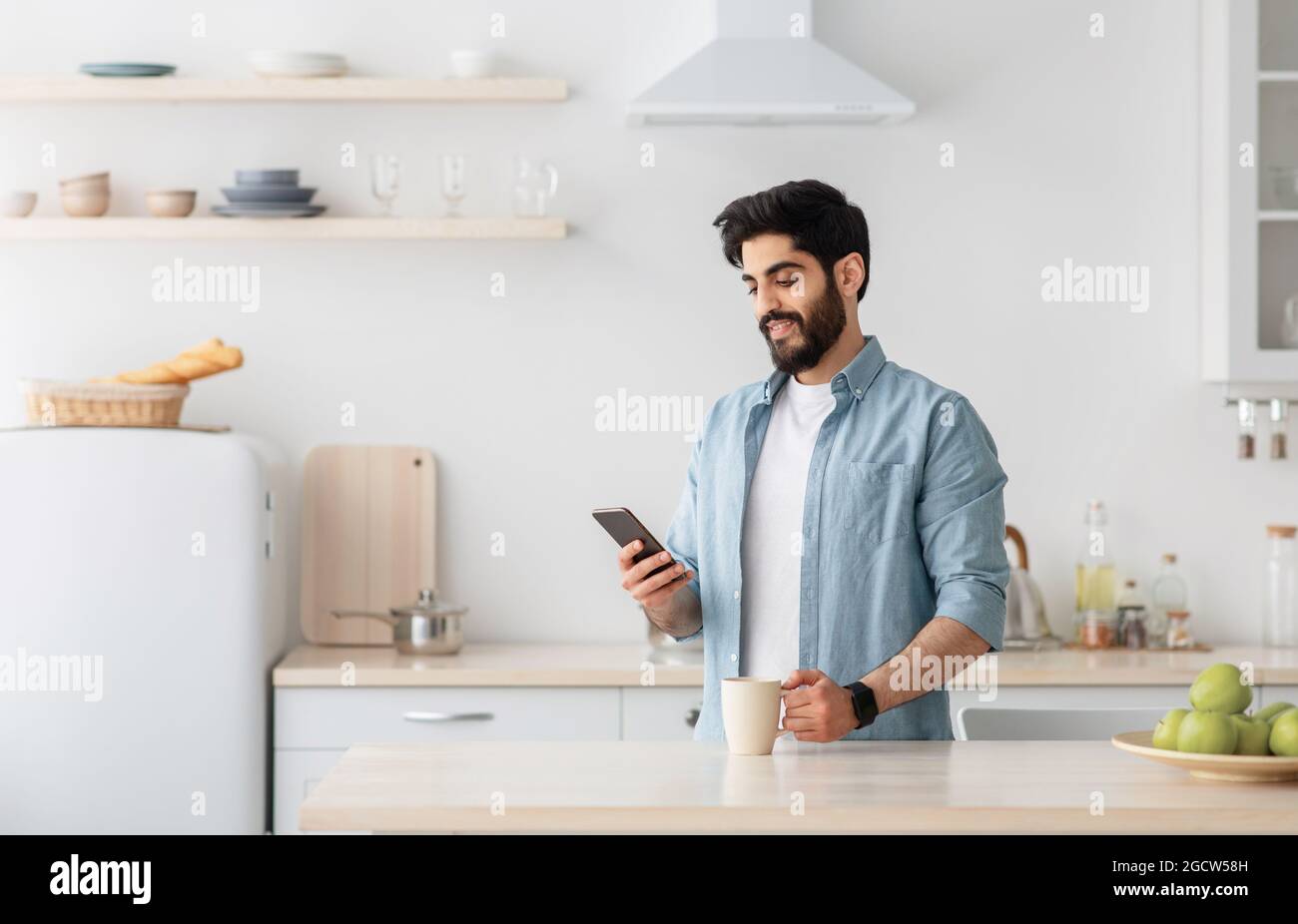 Happy young arab man using smartphone and drinking morning coffee in kitchen, empty space Stock Photo