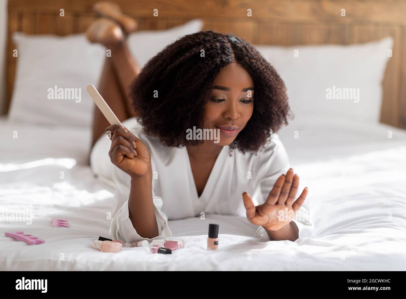 Pretty black woman lying on bed with nail file, doing manicure, taking