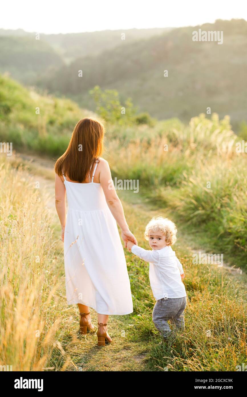 Elegant woman in white dress leads her son by the hand while walking in nature Stock Photo