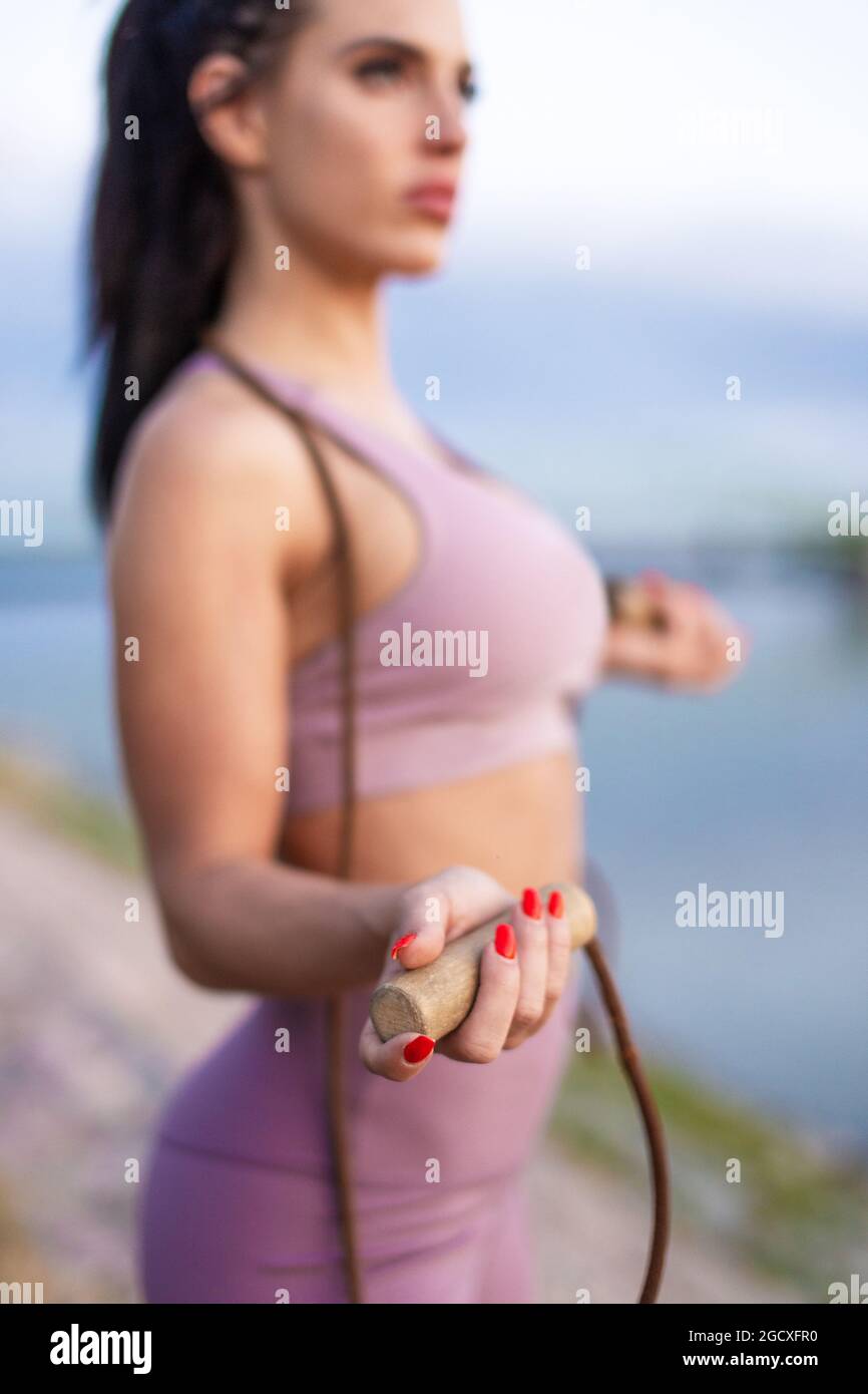 Young athlete woman holding jump rope at riverbank, cardio workout, depth of field Stock Photo