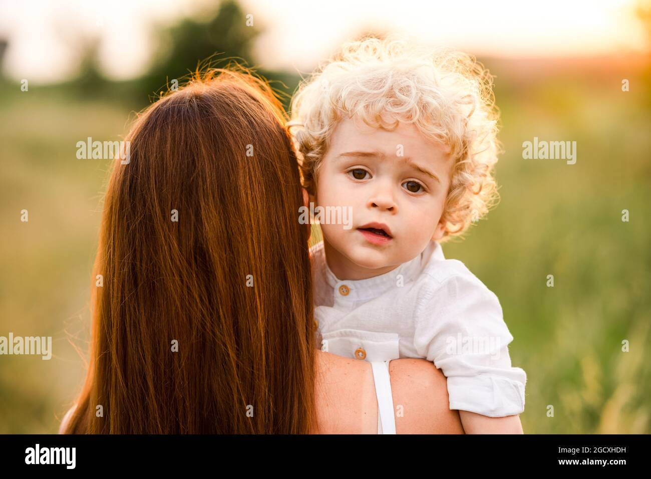 Portrait of a young curly-haired boy in his mother's arms while walking on a field at sunset Stock Photo