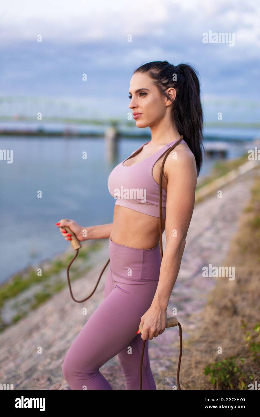 Young athlete woman holding jump rope at riverbank, cardio workout Stock Photo