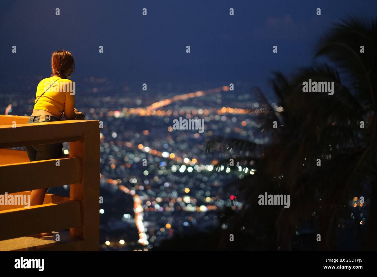 Young woman looks around night lights of a big city (Chiang Mai) while standing on a balcony high above Stock Photo