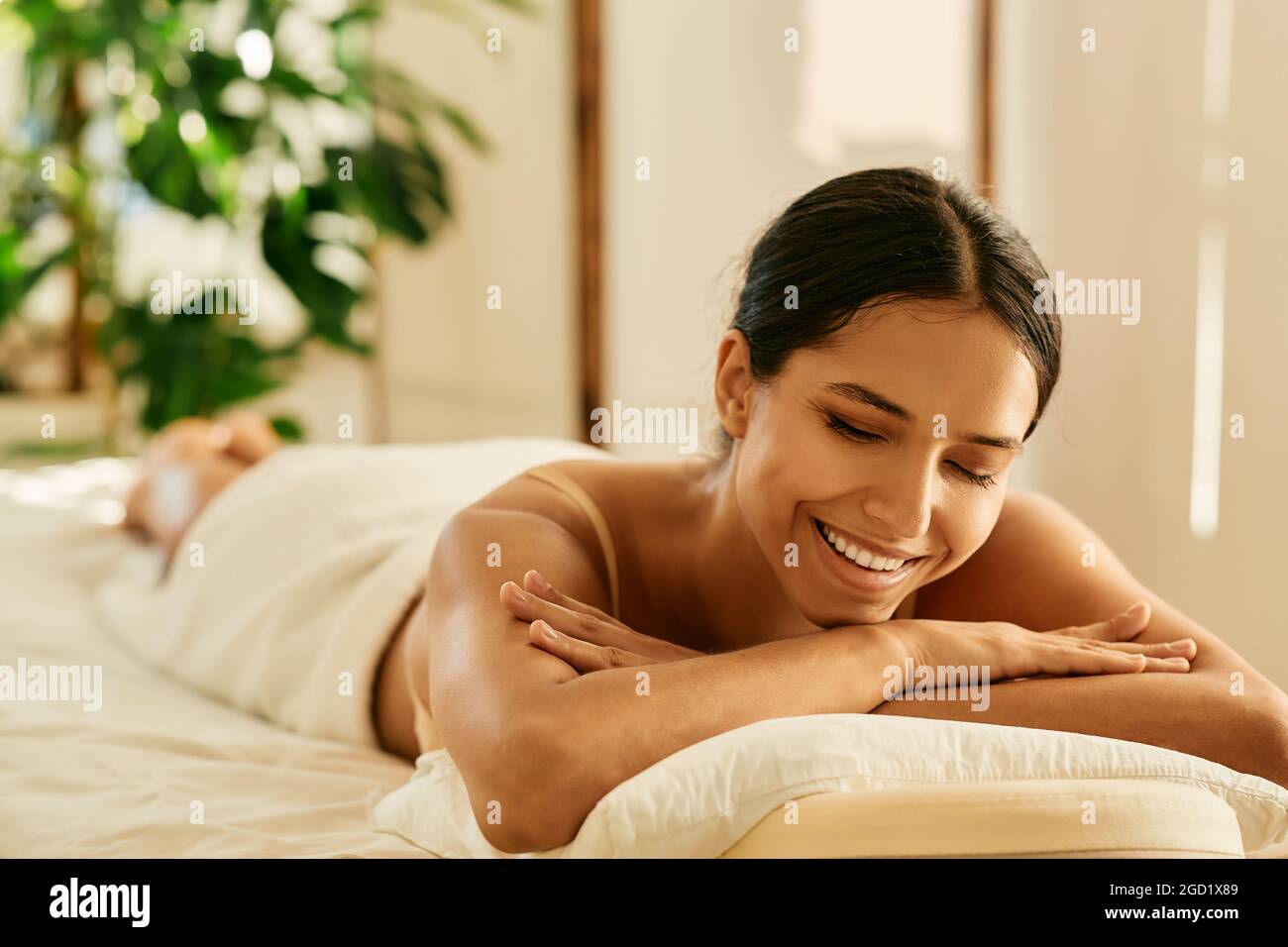 Beautiful mixed race woman with white smile lying down on massage table enjoys day spa at wellness salon Stock Photo
